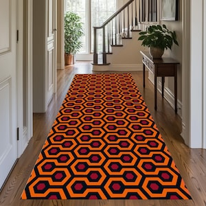 May include: A long, rectangular rug with a repeating geometric pattern of orange, black, and burgundy hexagons. The rug is placed in a hallway with wooden floors, white doors, and a staircase.