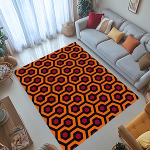 Puede incluir: Una alfombra rectangular con un patrón geométrico repetitivo de hexágonos naranjas, negros y burdeos. La alfombra está colocada sobre un suelo blanco en un salón, con un sofá y sillas visibles.