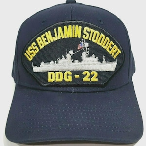 May include: Navy blue baseball cap featuring a patch with the text "USS BENJAMIN STODDERT" in yellow, arching over a white illustration of a ship. Below the ship is "DDG-22" in yellow. The cap has a curved brim and a button on top.