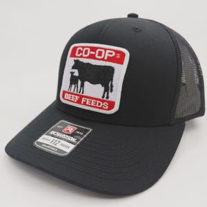 May include: Black trucker hat with a mesh back and a curved brim. The front features a patch with the text "CO-OP" above a silhouette of a cow and calf, with "BEEF FEEDS" below. The hat is adjustable.
