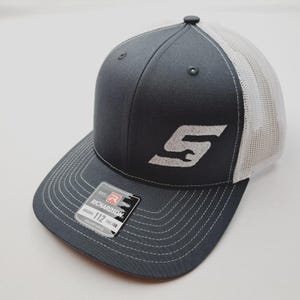 May include: A gray trucker hat with a white mesh back and a curved brim. The hat features a white embroidered logo and stitching. A tag on the brim reads "RICHARDSON 112 TRUCKER ADJUSTABLE."