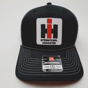 May include: Black and white trucker hat with the "International Harvester" logo on the front. The logo is a white square with a black and red design. The hat has a curved brim and a white mesh back. The tag reads "Richardson 112 Trucker".