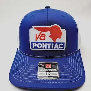 May include: A blue and white Richardson 112 Trucker hat with a stitched V8 Pontiac logo patch. The hat features a curved brim, white mesh back, and adjustable snap closure. The patch includes the text "V8 PONTIAC" and a red graphic.