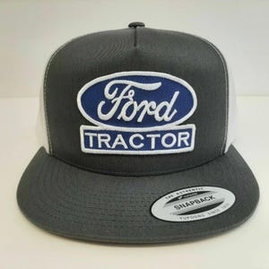 May include: A charcoal gray and white trucker hat featuring the Ford Tractor logo in blue and white. The hat has a flat brim and a snapback closure. The tag reads "The Authentic Yupoong Snapback Since 1974."