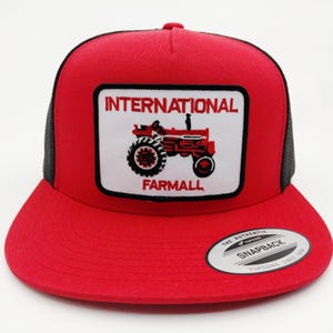 May include: A red snapback hat with a black mesh back and a flat brim. The front features a white patch with the words "INTERNATIONAL FARMALL" above a red tractor illustration. The hat has a curved brim and a snap closure.