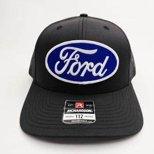 May include: A black trucker hat featuring a prominent blue and white oval Ford logo patch on the front. The cap includes a black mesh back and a black brim with a rectangular sticker that reads Richardson Est. 1970 Original 112 Trucker Adjustable.