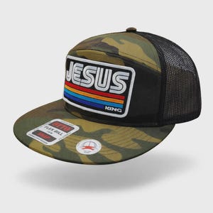 May include: Camouflage baseball cap with a black mesh back. The front features a patch with the word "JESUS" in white block letters above a rainbow stripe design. The word "KING" is below the stripes.