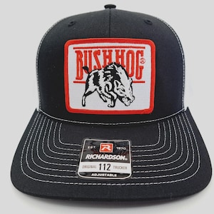 May include: Black trucker hat with a white mesh back and a square patch featuring the text "BUSH HOG" above a black and white illustration of a hog. The hat has a curved brim with white stitching and a "RICHARDSON" tag.