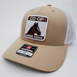 May include: Tan and white trucker hat with a patch that reads "CO-OP HORSE FEEDS" and features a brown horse head. The hat has a curved brim and a white mesh back. The Richardson brand label is visible.