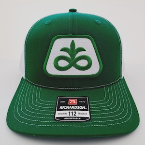May include: Green and white trucker hat with a white patch featuring a green design. The hat has a curved brim and a mesh back. The tag reads "RICHARDSON ORIGINAL 112 TRUCKER ADJUSTABLE."