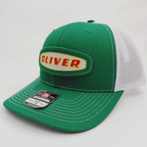 May include: Green and white trucker hat with the name "OLIVER" in red letters on a yellow and green patch. The hat has a curved brim and a white mesh back. The Richardson brand tag is visible.