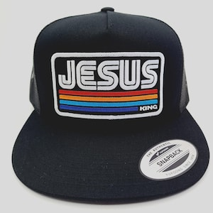 May include: Black trucker hat with a flat brim. The front features a rectangular patch with the word "JESUS" in white, outlined in black, above horizontal stripes of red, orange, blue, and a thin blue stripe. The word "KING" is below the stripes.