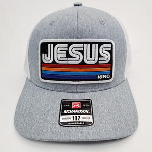 May include: A gray trucker hat with a white mesh back and a curved brim. The front patch reads "JESUS KING" in white letters over a striped design. The hat includes a Richardson 112 Trucker tag.