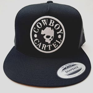 May include: Black trucker hat with a flat brim and a white embroidered patch. The patch features the words "COWBOY CARTEL" around a skull design. The hat has a mesh back and a snapback closure.
