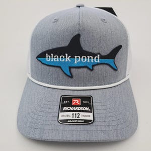 May include: Gray trucker hat with a white mesh back and a black and blue shark graphic that reads "black pond." The hat has a white rope detail and a Richardson patch.