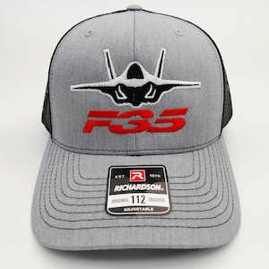 May include: A grey trucker hat with a black mesh back and a structured front panel. The front features an embroidered black and white F-35 fighter jet graphic above the red text F35. A black Richardson 112 adjustable sticker is on the brim.