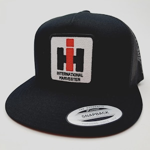 May include: Black snapback hat with a white patch featuring the International Harvester logo in black and red. The hat has a curved brim and a mesh back. The hat has a Snapback closure.