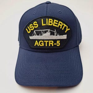 May include: Navy blue baseball cap with a curved brim. The cap features a patch with the text "USS LIBERTY AGTR-5" in yellow and white, with an image of a ship. The cap is made of a durable fabric.