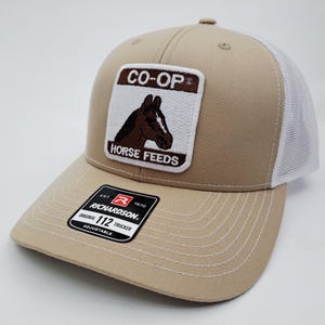 May include: Tan and white trucker hat with a patch featuring a brown horse head and the words "CO-OP HORSE FEEDS". The hat has a curved brim and an adjustable strap. The Richardson brand label is visible.