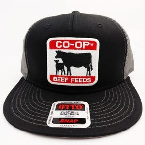 May include: Black and gray snapback hat with a flat bill. The front features a patch with the text "CO-OP" above a silhouette of a cow and calf, with "BEEF FEEDS" below. The hat has a classic fit and snap closure.