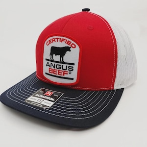 May include: A red, white, and navy blue trucker hat with a mesh back. The front features a patch with the words "CERTIFIED ANGUS BEEF" and a silhouette of a bull. The hat is adjustable.