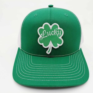 May include: Green and white baseball cap featuring a shamrock with the word "Lucky" embroidered in white. The cap has a green brim and crown, with a white mesh back. The stitching is white.