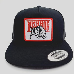 May include: Black trucker hat with a mesh back and a flat brim. The front features a rectangular patch with the text "BUSH HOG" above a black and white illustration of a hog. The hat has a snapback closure.