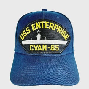 May include: A navy blue baseball cap featuring a patch with the text "USS ENTERPRISE CVAN-65" in yellow and white. The patch depicts a stylized image of an aircraft carrier. The cap has a curved brim and a six-panel design.