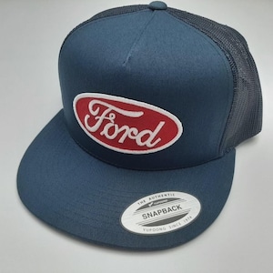 May include: A navy blue trucker hat with a curved brim and mesh back. The front features a red oval patch with the white embroidered word "Ford". A white tag on the brim reads "The Authentic Snapback Yupoong Since 1974".