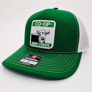 May include: Green and white trucker hat with a square patch featuring a cow illustration and the words "CO-OP" and "BEEF FEEDS." The hat has a curved brim and a white mesh back. The Richardson 112 Trucker hat is adjustable.