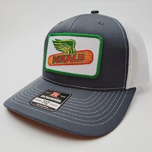 May include: A charcoal gray trucker hat with a white mesh back. The front features a rectangular patch with the word "DEKALB" in red and gold, a green winged design, and a green border. The hat has a curved brim and a "Richardson" tag.