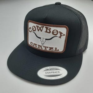May include: Black snapback trucker hat with a mesh back and a flat brim. The front features a rectangular patch with the words "COWBOY CARTEL" and a bull skull graphic. The hat also has a "Yupoong" label.