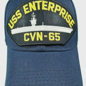 May include: Navy blue baseball cap featuring a patch with the text "USS ENTERPRISE CVN-65" in yellow lettering. The patch also includes a white and black graphic of a ship. The cap is made of a textured fabric.