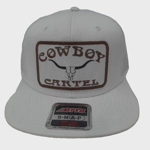 May include: White snapback hat with a brown patch featuring the text "COWBOY CARTEL" and a longhorn skull graphic. The hat has a curved brim and a mesh back. The hat is one size fits most.