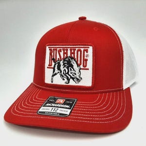 May include: Red and white trucker hat with a patch featuring the text "RUSH HOG" above a black and white illustration of a boar. The hat has a curved brim, white mesh back, and an adjustable strap. The Richardson brand tag is visible.