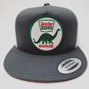 May include: A gray and white trucker hat featuring a circular patch with the Sinclair Dino logo. The patch includes a green dinosaur, the words "Sinclair DINO" and "GASOLINE" in red. The hat has a flat brim and a snapback closure.