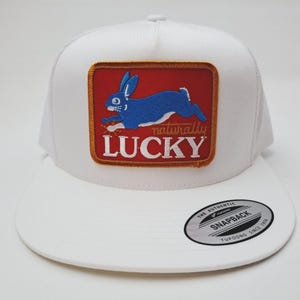 May include: White trucker hat with a patch featuring a blue rabbit leaping across a red background. The patch reads "naturally LUCKY" in white lettering. The hat has a curved brim and a snapback closure.