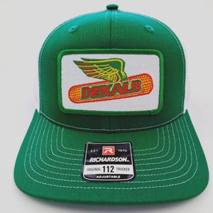 May include: Green and white trucker hat with a patch featuring the word "DEKALB" in red and yellow, with a winged design. The hat has a curved brim and an adjustable strap. The tag reads "RICHARDSON ORIGINAL 112 TRUCKER ADJUSTABLE".