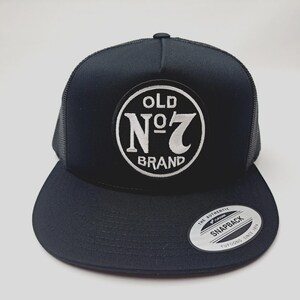 May include: Black trucker hat with a curved brim and mesh back. The front features a white circular patch with the text "OLD N°7 BRAND". A small, silver sticker with "SNAPBACK" is on the brim.
