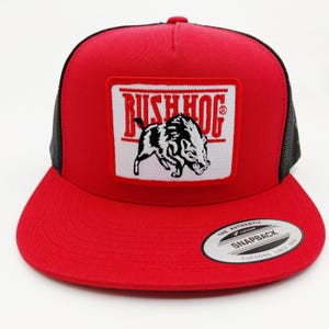 May include: A red trucker hat with a black mesh back and a flat brim. The front features a white patch with the word "BUSH HOG" and a black and white illustration of a wild boar. The hat has a snapback closure.