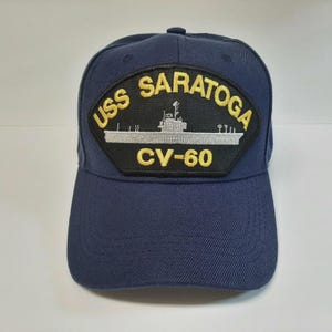 May include: Navy blue baseball cap with a patch that reads "USS SARATOGA CV-60". The patch features a silver illustration of a ship. The cap has a curved brim and a button on top.