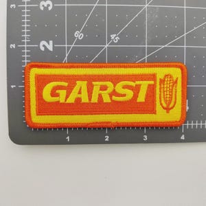 May include: Rectangular orange and yellow embroidered patch with the word "GARST" in yellow block letters. A yellow corn graphic is on the right side. The patch is on a gray surface with measurement markings.