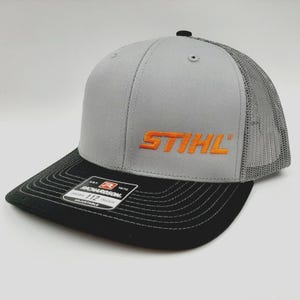 May include: Gray and black baseball cap with the word "STIHL" embroidered in orange. The cap has a black brim and a mesh back. The Richardson 112 adjustable hat is a trucker style.