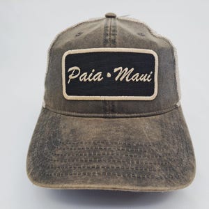 May include: A distressed brown baseball cap with a mesh back. The front features a black rectangular patch with the words "Paia • Maui" embroidered in white. The cap has a curved brim and a button on top.