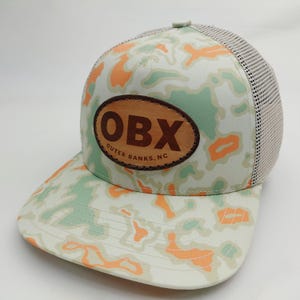 May include: A baseball cap featuring a light green, white, and orange camouflage pattern. The front panel displays an oval-shaped brown leather patch with the text OBX and OUTER BANKS, NC embossed in dark brown lettering. The back panels are mesh.
