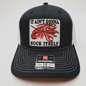 May include: Black and white trucker hat with a patch featuring a cartoon crawfish and the text "IT AIN'T GONNA SUCK ITSELF." The hat has a curved brim and white mesh back. The Richardson brand tag is visible.