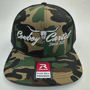 May include: A camouflage flat bill snapback hat with a black mesh back. The hat features the words "Cowboy Cartel" in white script, along with a white bull skull graphic and "Since 2021". A Richardson tag is visible.