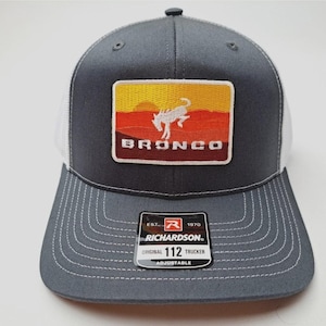 May include: A gray and white trucker hat with a rectangular patch featuring a white horse silhouette against a sunset backdrop. The word "BRONCO" is below the horse. The hat has a curved brim and a Richardson tag.