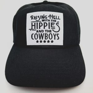 May include: Black baseball cap with a white square patch. The patch features the text "RAISING HELL WITH THE HIPPIES AND THE COWBOYS" in a stylized font, with five stars below the text. The cap has a curved brim.