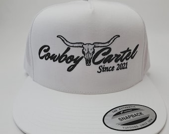 May include: White trucker hat with a flat brim. The hat features the text "Cowboy Cartel Since 2021" in black script with a longhorn skull graphic. A "Snapback" sticker is on the brim.
